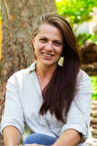 Woman with long brown hair and a white shirt and jeans sitting with her legs crossed and smiling at the camera. There is a tree and some greenery in the background