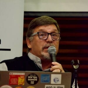 An image of Gerado, a man with short dark hair, goatee and glasses, speaking into a microphone at a conference.