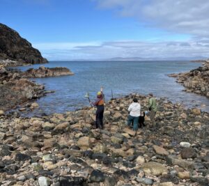Three people standing in Camas Bay throwing rocks at the Camas bell