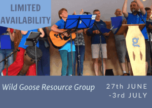 Group of people singing on a stage. One person has a guitar and one has their hands clapping in the air. There is a row of people standing holding blue open files behind them, they are singing.