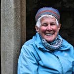 Woman wearing a colouful hat and blue coat smiling at camera, she's leaning against one of the pillars in the cloisters at Iona Abbey