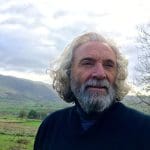 A man with grey hair and beard with hills in the background.