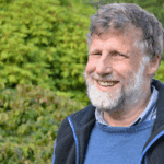 Man with navy fleece and blue jumper smiling at camera, he's standing in front of lots of greenery