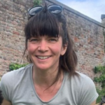 Woman with sunglasses on her long brown hair wearing a beige t-shirt. She is smiling.