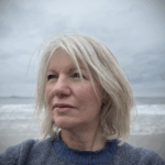 Woman with blonde hair wearing a grey jumper standing in front of the sea/