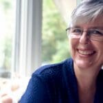 woman with glasses and navy top smiling at camera, she's leaning forward and has a warm friendly face