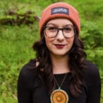 Woman with black top, black hair and an orange wooly hat stands in front of greenery