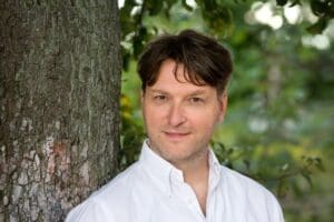 A man with brown hair and white shirt standing beside a tree - rewild your spirit