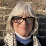 Woman with grey bobbed hair wearing black glasses, a white polo neck and a cream scarf with grey dots on it.