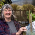 Margaret Elphinstone Photo, she is wearing a red and navy checked shirt and is standing in a garden, there is a small growing tunnel behind her to the right.