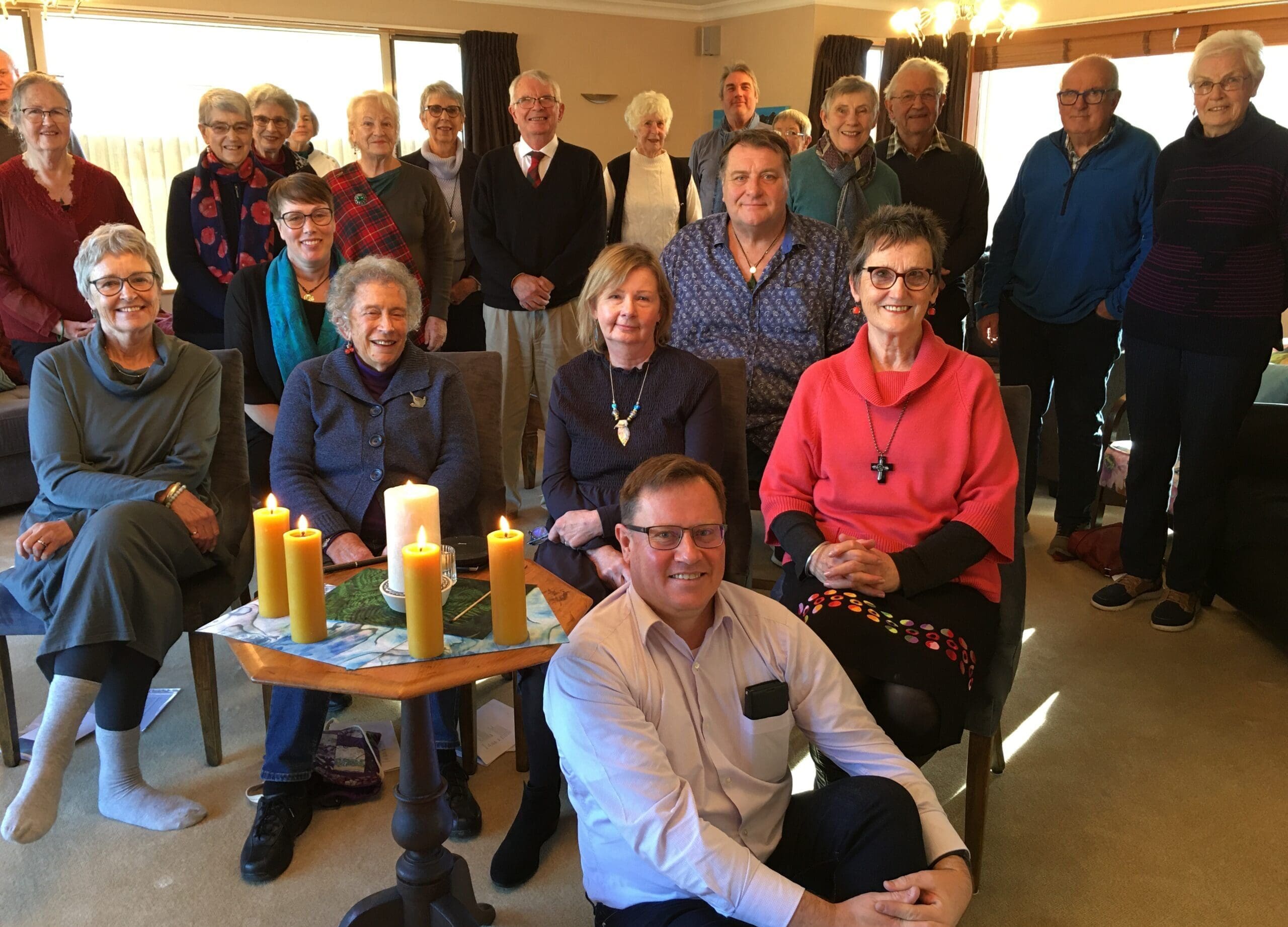 A large group of people sitting in a living room with five candles lit in the middle of the room