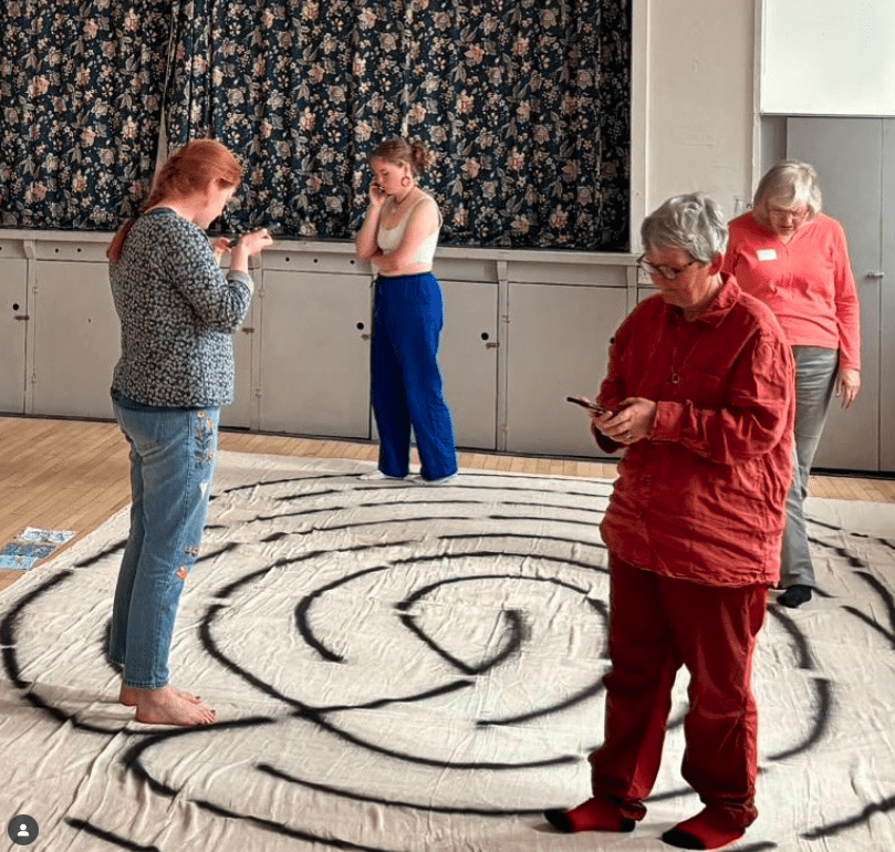 Four people walking the fingerprint labyrinth. They're spaced apart from each other. Two of them are using their phones as they walk the labyrinth to help them engage with the associated materials.