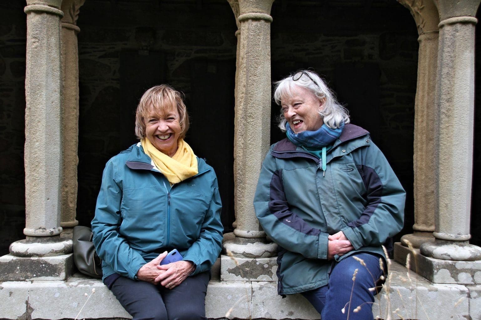 Two women sat in the cloisters of Iona Abbey. The one on the right is laughing as she looks at the one on the left.The one on the left is wearing a yellow scarf. They are both wearing a blue/ green coat.