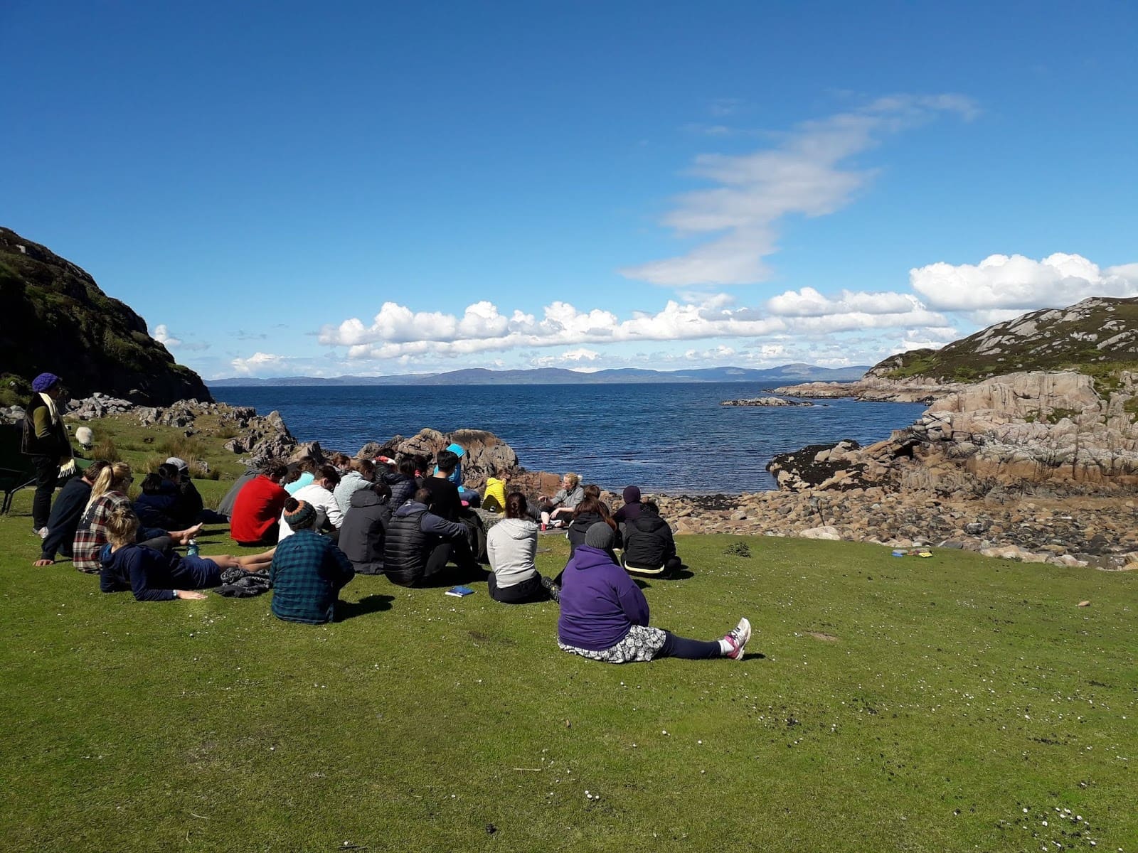 Group of people sitting on grass looking out to sea.